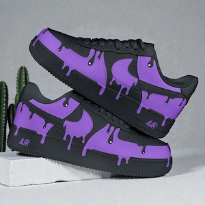 Può includere: Un paio di sneakers nere e viola con un design a goccia. Le scarpe hanno una base nera con dettagli viola e un effetto vernice gocciolante. La parola "AIR" è stampata sul lato. Le scarpe sono su una superficie bianca.
