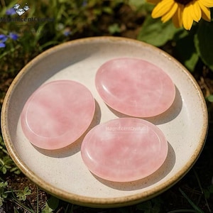 May include: Three oval rose quartz stones rest in a beige ceramic bowl. The stones are a soft pink color with subtle variations in tone. The bowl sits on a bed of green grass, with a yellow sunflower and blue flowers in the background. The text "MagnificentCrystal" is visible.