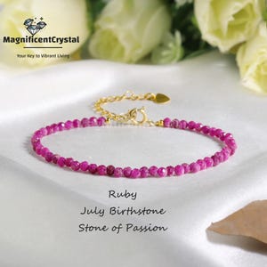 May include: A delicate ruby bracelet with a gold chain and clasp. The bracelet is made of small, faceted ruby beads. Text on the image reads "Ruby, July Birthstone, Stone of Passion" and "MagnificentCrystal, Your Key to Vibrant Living."