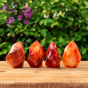 May include: Four carnelian crystal points in various shades of orange and red, arranged in a row on a wooden surface. The crystals are smooth and polished, with a natural, organic shape.