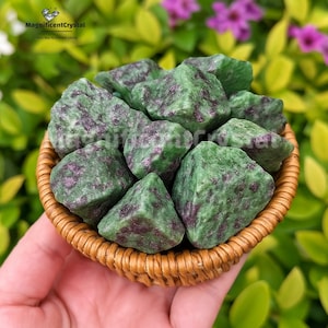 May include: A woven basket filled with rough-cut green zoisite stones with dark red inclusions. The stones are held in a small, round, woven basket. The background is blurred, showing green foliage and purple flowers.
