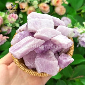 May include: A woven basket filled with raw, light purple crystals. The crystals are irregularly shaped and have a rough texture. The background features blurred flowers and greenery. The text "Magnificent Crystal" is visible.