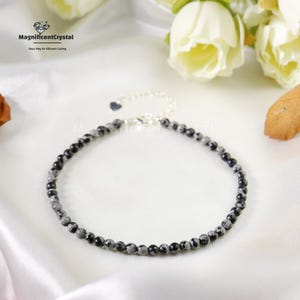 May include: Ankle bracelet featuring small, round, black and gray speckled beads strung on a silver chain. The bracelet has a heart-shaped charm and a clasp closure. The background includes white flowers and the text "MagnificentCrystal".