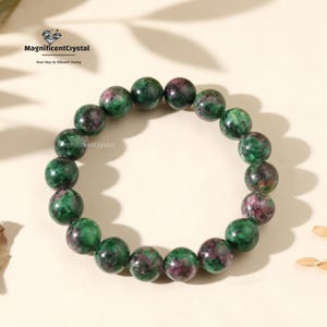 May include: A beaded bracelet featuring round, polished gemstones in shades of green and maroon. The bracelet is displayed on a light-colored surface, with the text "MagnificentCrystal" and "Your Key to Vibrant Living" visible in the upper left corner.