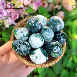 May include: A collection of polished moss agate spheres in a woven basket. The spheres are primarily white and green, with intricate patterns. The basket is held against a backdrop of green foliage and pink flowers. Text on the image reads "MagnificentCrystal" and "Your Key to Vibrant Living."
