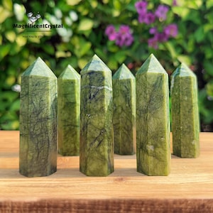 Könnte beinhalten: Sieben grüne Serpentin-Kristallspitzen, auch bekannt als grüner Jade, sind in einer Reihe auf einer Holzoberfläche angeordnet. Die Spitzen sind alle ungefähr gleich groß und geformt und haben eine glatte, polierte Oberfläche.