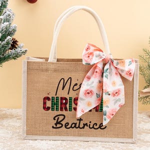 May include: A burlap tote bag with white handles and a floral bow. The bag has the words "Merry Christmas Beatrice" printed on the front in black and red plaid lettering. A small Christmas tree is on the left.