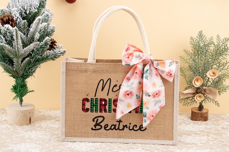 May include: A burlap tote bag with white handles and a floral bow. The bag has the words "Merry Christmas Beatrice" printed on the front in black and red plaid lettering. A small Christmas tree is on the left.