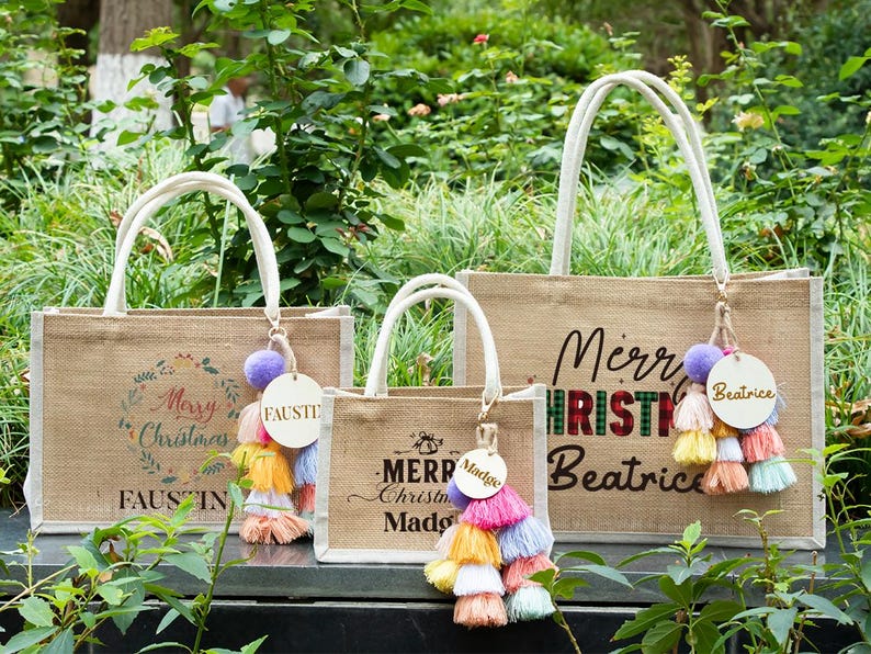 May include: Three burlap tote bags with white handles and festive Christmas designs. Each bag features a colorful tassel and a personalized wooden tag. The bags have the words "Merry Christmas" and names printed on them.