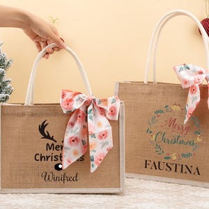 May include: Two rectangular burlap tote bags with white handles and floral bows. One bag has a deer antler graphic and the text "Merry Christmas Winifred". The other bag has a floral wreath and the text "Merry Christmas Faustina".