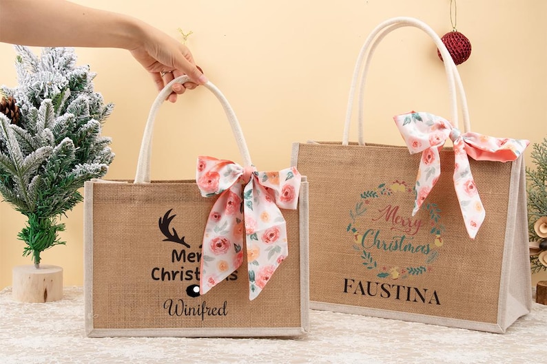 May include: Two rectangular burlap tote bags with white handles and floral bows. One bag has a deer antler graphic and the text "Merry Christmas Winifred". The other bag has a floral wreath and the text "Merry Christmas Faustina".