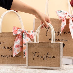 May include: Three brown burlap tote bags with white handles and floral bows. The bags have the name "Faustina" in black script, with "Merry Christmas" on the largest bag.