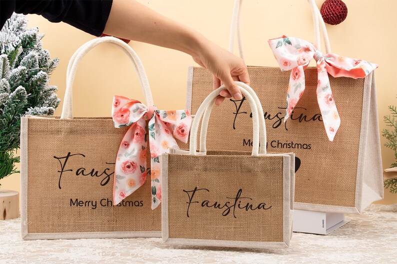 May include: Three brown burlap tote bags with white handles and floral bows. The bags have the name "Faustina" in black script, with "Merry Christmas" on the largest bag.