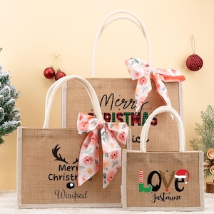 May include: Three burlap tote bags with white handles and floral bows. The largest bag says "Merry Christmas" and the middle bag says "Merry Christmas Winifred". The smallest bag says "Love Jasmine" with a Santa hat.