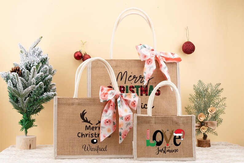 May include: Three burlap tote bags with white handles and floral bows. The largest bag says "Merry Christmas" and the middle bag says "Merry Christmas Winifred". The smallest bag says "Love Jasmine" with a Santa hat.