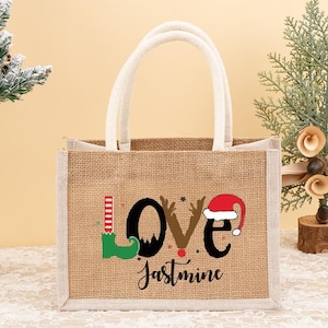 May include: A burlap tote bag with white handles and trim. The bag features the word "LOVE" with a Christmas theme, including an elf shoe, reindeer antlers, and a Santa hat. The name "Jastmine" is written below.