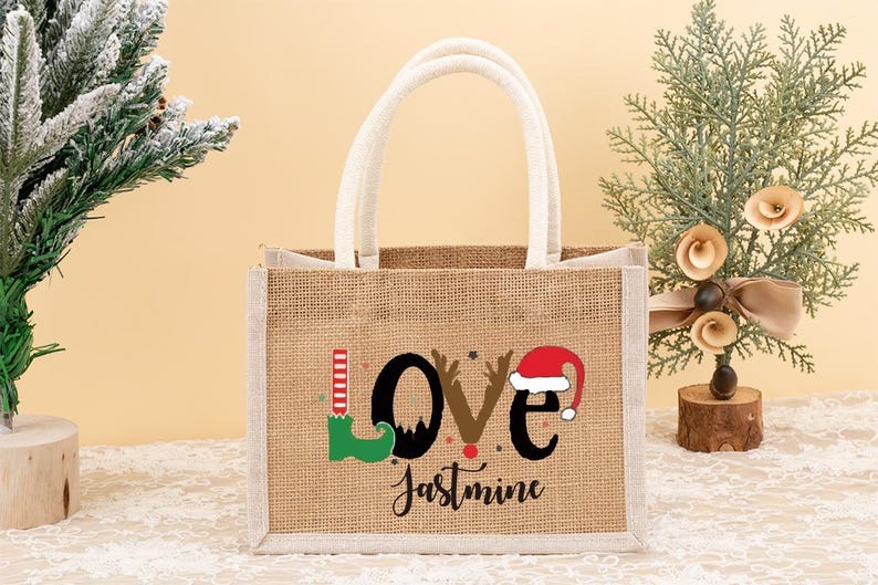 May include: A burlap tote bag with white handles and trim. The bag features the word "LOVE" with a Christmas theme, including an elf shoe, reindeer antlers, and a Santa hat. The name "Jastmine" is written below.