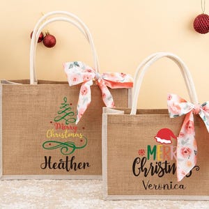 May include: Two burlap tote bags with white handles and floral bows. One bag has a Christmas tree graphic and the name "Heather". The other bag has a Santa hat and the name "Veronica". Both bags have "Merry Christmas" text.