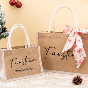 May include: Two burlap tote bags with white handles. The larger bag has a floral ribbon tied to the handle. Both bags have the name "Faustina" printed on them. The smaller bag also has "Merry Christmas" printed on it.