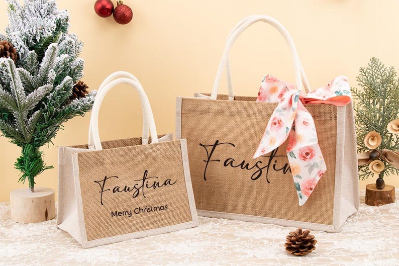 May include: Two burlap tote bags with white handles. The larger bag has a floral ribbon tied to the handle. Both bags have the name "Faustina" printed on them. The smaller bag also has "Merry Christmas" printed on it.