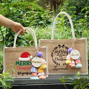May include: Two burlap tote bags with white handles. One bag has a Santa hat graphic and the words "Merry Christmas Olympia". The other bag has a wreath graphic and the words "Merry Christmas Laurinda". Each bag has a tassel charm.