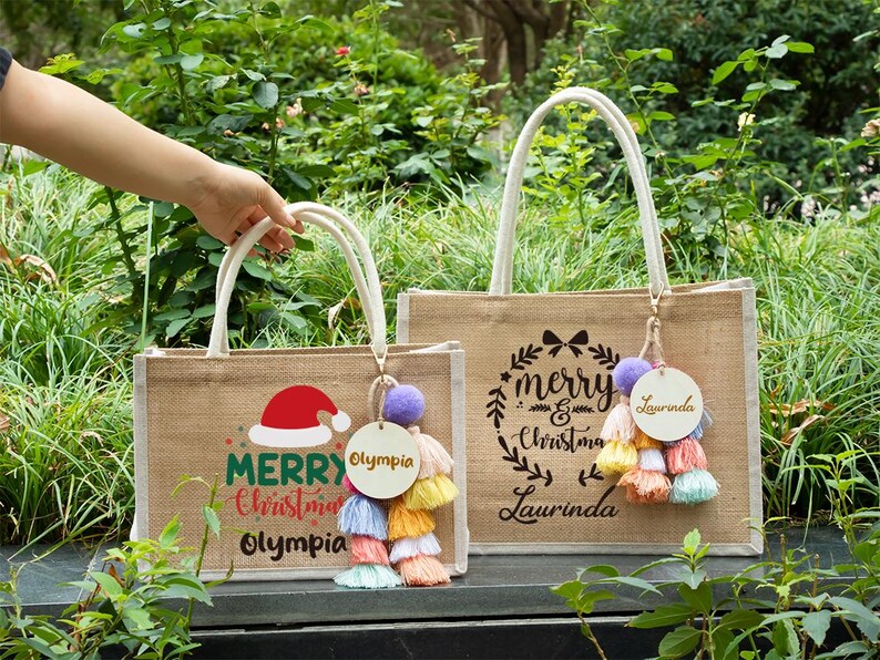 May include: Two burlap tote bags with white handles. One bag has a Santa hat graphic and the words "Merry Christmas Olympia". The other bag has a wreath graphic and the words "Merry Christmas Laurinda". Each bag has a tassel charm.