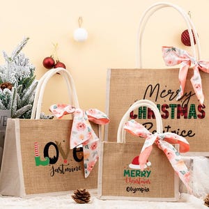 May include: Three burlap tote bags with white handles and floral bows. The bags have Christmas-themed text: "Merry Christmas Patrice", "Love Jasmine", and "Merry Christmas Olympia". A book and decorative pine tree are in the background.