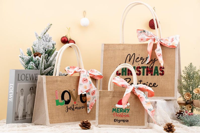 May include: Three burlap tote bags with white handles and floral bows. The bags have Christmas-themed text: "Merry Christmas Patrice", "Love Jasmine", and "Merry Christmas Olympia". A book and decorative pine tree are in the background.