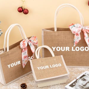 Peut inclure: Trois sacs fourre-tout en toile de jute avec des poignées blanches et des rubans à motifs floraux. Le texte "YOUR LOGO" est imprimé sur les sacs. Un petit sapin de Noël et des décorations sont en arrière-plan.