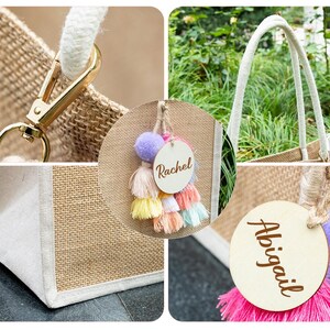 May include: A natural jute tote bag with white trim and rope handles. The bag features a gold-colored clasp and a decorative tassel with a wooden tag that says "Rachel".