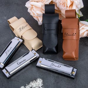 Puede incluir: Tres armónicas con nombres personalizados, "Katrisha", "Queenie" y "Blanche", se muestran con sus estuches de cuero a juego. Los estuches son beige, negro y marrón.