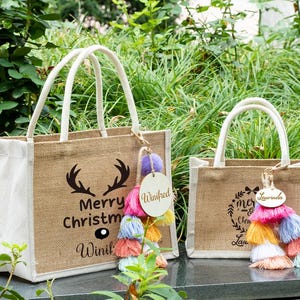 May include: Two burlap tote bags with white handles. One bag has a black antler design and the text "Merry Christmas Winifrd". The other bag has a wreath design and the text "Merry Christmas Laurinda". Both bags have colorful tassel keychains.