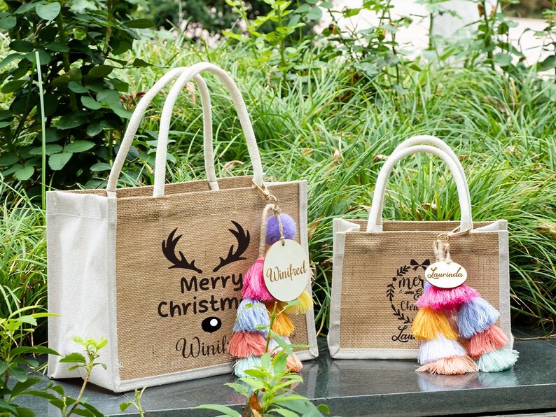 May include: Two burlap tote bags with white handles. One bag has a black antler design and the text "Merry Christmas Winifrd". The other bag has a wreath design and the text "Merry Christmas Laurinda". Both bags have colorful tassel keychains.