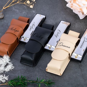 May include: Five personalized harmonicas in leather cases. The harmonicas are silver with black lettering. The cases are brown, black, beige, and black. The names on the harmonicas are Helga, Vicky, Ingrid, Deanna, and Deanna.