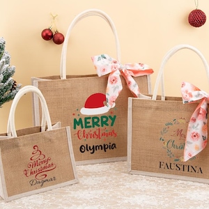 May include: Three burlap tote bags with white handles and festive Christmas designs. One bag features a Santa hat and the words "Merry Christmas Olympia." Another has a floral bow and the name "Faustina."