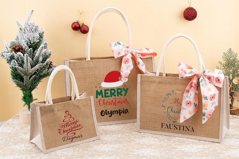 May include: Three burlap tote bags with white handles and festive Christmas designs. One bag features a Santa hat and the words "Merry Christmas Olympia." Another has a floral bow and the name "Faustina."