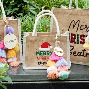 May include: Jute tote bags with white handles, featuring Christmas-themed designs and personalized wooden tags. The bags have colorful tassel keychains. One bag says "Merry Christmas Olympia", another says "Faustina", and another says "Beatrice".