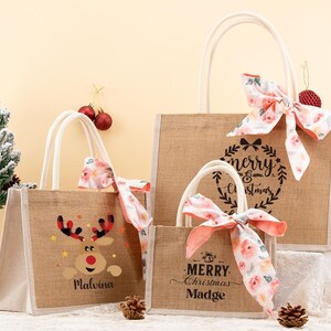 May include: Three burlap tote bags with white handles and floral bows. One bag features a reindeer design, another says "Merry Christmas Madge", and the largest says "Merry Christmas". Christmas-themed gift bags.