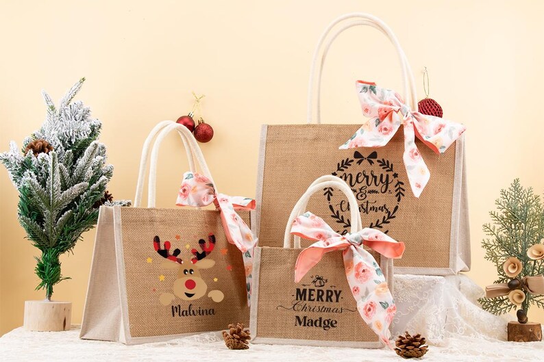 May include: Three burlap tote bags with white handles and floral bows. One bag features a reindeer design, another says "Merry Christmas Madge", and the largest says "Merry Christmas". Christmas-themed gift bags.