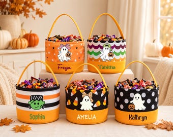 May include: Five cylindrical fabric Halloween candy buckets with yellow handles, each filled with assorted candy. The buckets feature personalized names: Freya, Tabitha, Sophia, Amelia, and Kathryn, alongside various ghost, pumpkin, and monster graphics.