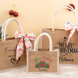 May include: Three burlap tote bags with white handles and floral bows. The bags have holiday-themed text, including "Merry Christmas" and names. The smallest bag has a gift graphic. The bags are a festive holiday gift.