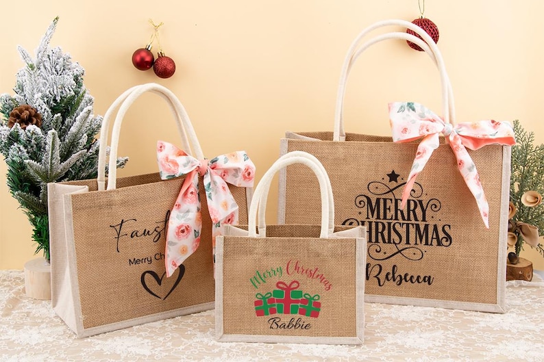 May include: Three burlap tote bags with white handles and floral bows. The bags have holiday-themed text, including "Merry Christmas" and names. The smallest bag has a gift graphic. The bags are a festive holiday gift.