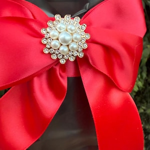 May include: A bright red satin bow with a gold and pearl brooch. The brooch is round with a cluster of pearls in the center, surrounded by smaller clear crystals. The bow is tied around a black object.