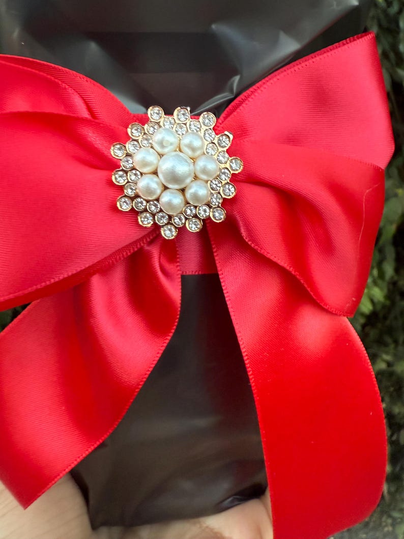 May include: A bright red satin bow with a gold and pearl brooch. The brooch is round with a cluster of pearls in the center, surrounded by smaller clear crystals. The bow is tied around a black object.