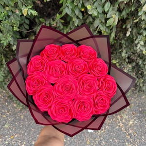 May include: A bouquet of vibrant pink roses, artfully arranged and wrapped in a dark burgundy paper. The bouquet is held against a backdrop of green foliage and a gray surface.