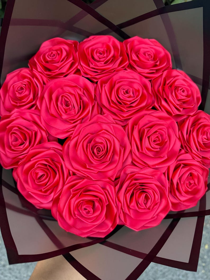 May include: A bouquet of vibrant pink fabric roses, artfully arranged. The roses are tightly clustered, showcasing their detailed petal construction. The bouquet is wrapped in a dark, patterned cellophane, creating a striking contrast.