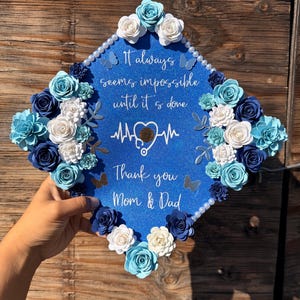 May include: A blue graduation cap decorated with paper flowers in shades of blue and white. The cap has the text "It always seems impossible until it is done" and "Thank you Mom & Dad".