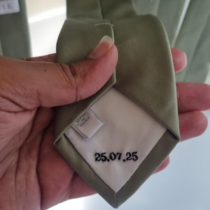 Può includere: Cravatta verde oliva con interno bianco. La data "25.07.25" è ricamata in nero. L'etichetta recita "MADE IN CHINA".