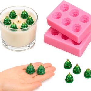 May include: A glass candle containing six small green Christmas tree-shaped wax embeds with gold-colored accents, two rectangular pink silicone molds with six cavities each, and several loose green tree-shaped wax embeds.
