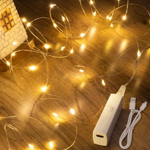 May include: A string of thin, flexible copper wire fairy lights emitting a warm yellow glow. The lights are connected to a rectangular white plastic battery pack with a USB charging cable attached, featuring a standard USB-A connector.